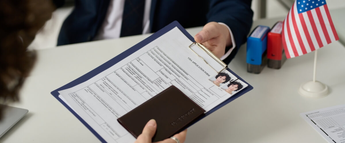 Hands holding L‑1A visa application form for Nigerian executives with U.S. flag in background.
