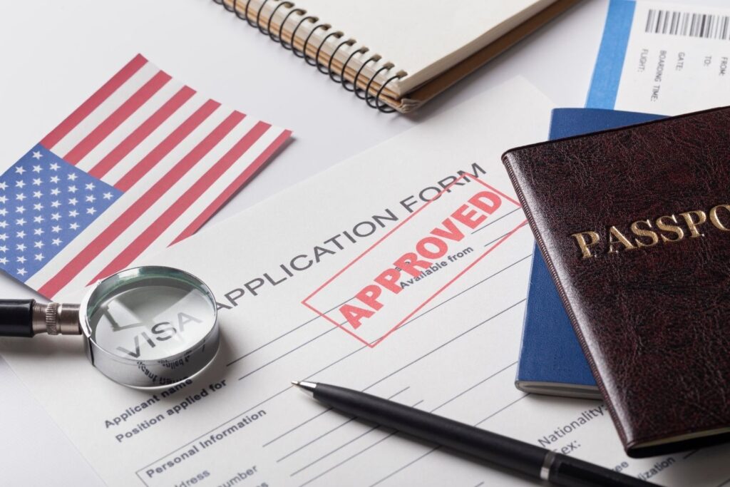 Close-up of a US visa application form stamped approved, a magnifying glass, passports, a pen, and a small American flag, symbolizing successful US visa approval.